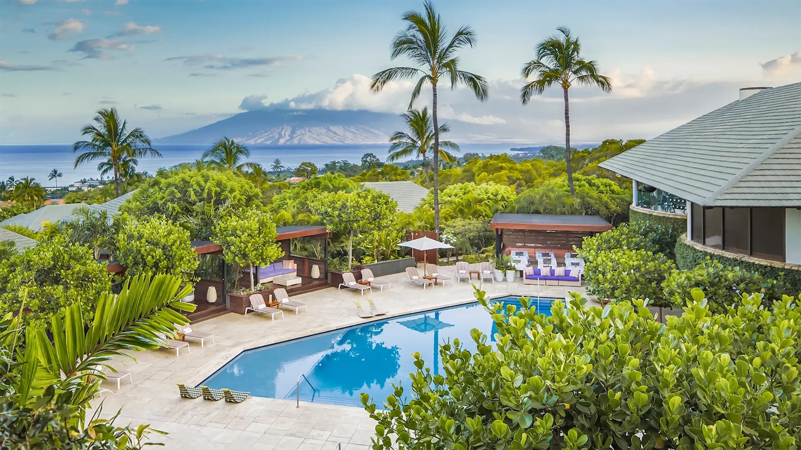 A scenic view of a resort in Hawaii featuring a swimming pool surrounded by lounge chairs and greenery. In the background, lush foliage leads to a clear blue ocean with a mountainous island rising in the distance under a lightly clouded sky, perfect for a romantic getaway.