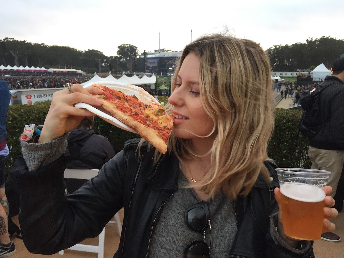 A woman with blonde hair wearing sunglasses on her shirt is holding a slice of pizza in one hand and a plastic cup of beer in the other. She appears to be taking a bite of the pizza. The background shows an outdoor event with people, tents, and trees.