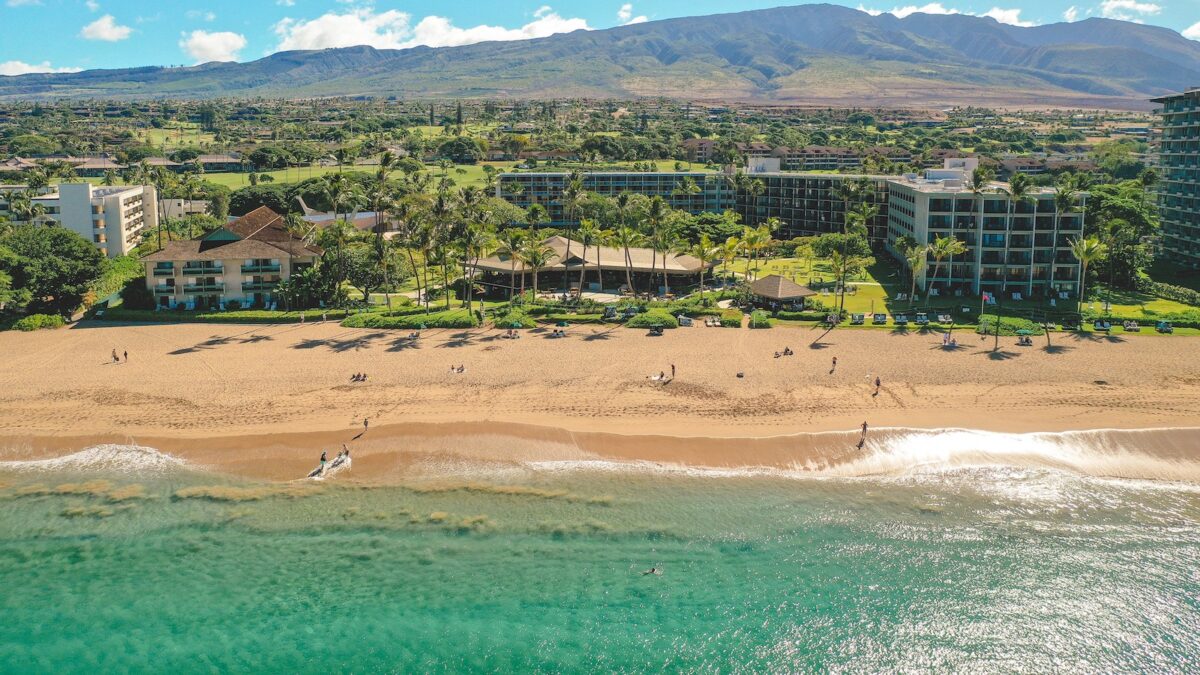 Aerial view of a sandy beachfront resort with several buildings surrounded by lush greenery and palm trees. Visitors walk along the beach and swim in the clear turquoise waters. Rolling mountains are visible in the background under a bright blue sky with scattered clouds.