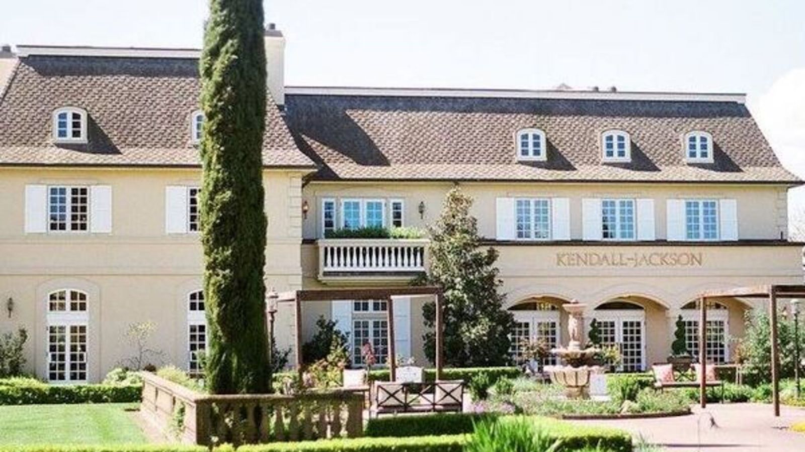 A large, elegant, cream-colored building with a brown roof and several windows with blue shutters. The entrance features arches and the name "Kendall-Jackson" displayed above. A fountain, greenery, and a tall tree enhance the landscape of one of the finest dog-friendly tasting rooms in Sonoma County.