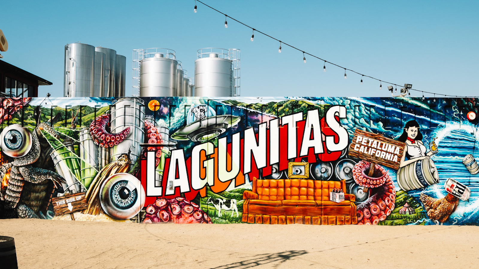 A vibrant mural featuring the word "LAGUNITAS" in large, bold letters on a colorful, eclectic background. The mural includes images of a couch, various beverages, water scenes, an octopus, and a diverse mix of objects—evoking the lively atmosphere of dog-friendly tasting rooms in Sonoma County. Above hang string lights and behind are metal tanks.