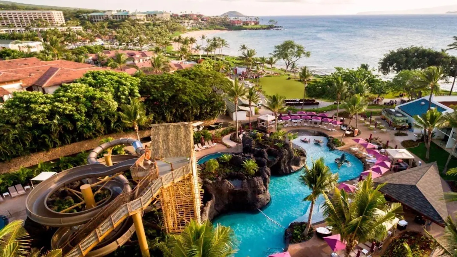 Aerial view of a tropical resort featuring a large swimming pool with a waterfall, a twisting waterslide, surrounding lush greenery, palm trees, sun loungers, and a beautiful ocean backdrop with the coastline visible.