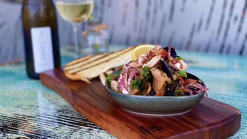 A bowl of mussels garnished with herbs and lemon sits on a wooden board, accompanied by toasted bread slices. A bottle of white wine and a glass filled with wine are in the background, placed on a table with a colorful, textured surface.