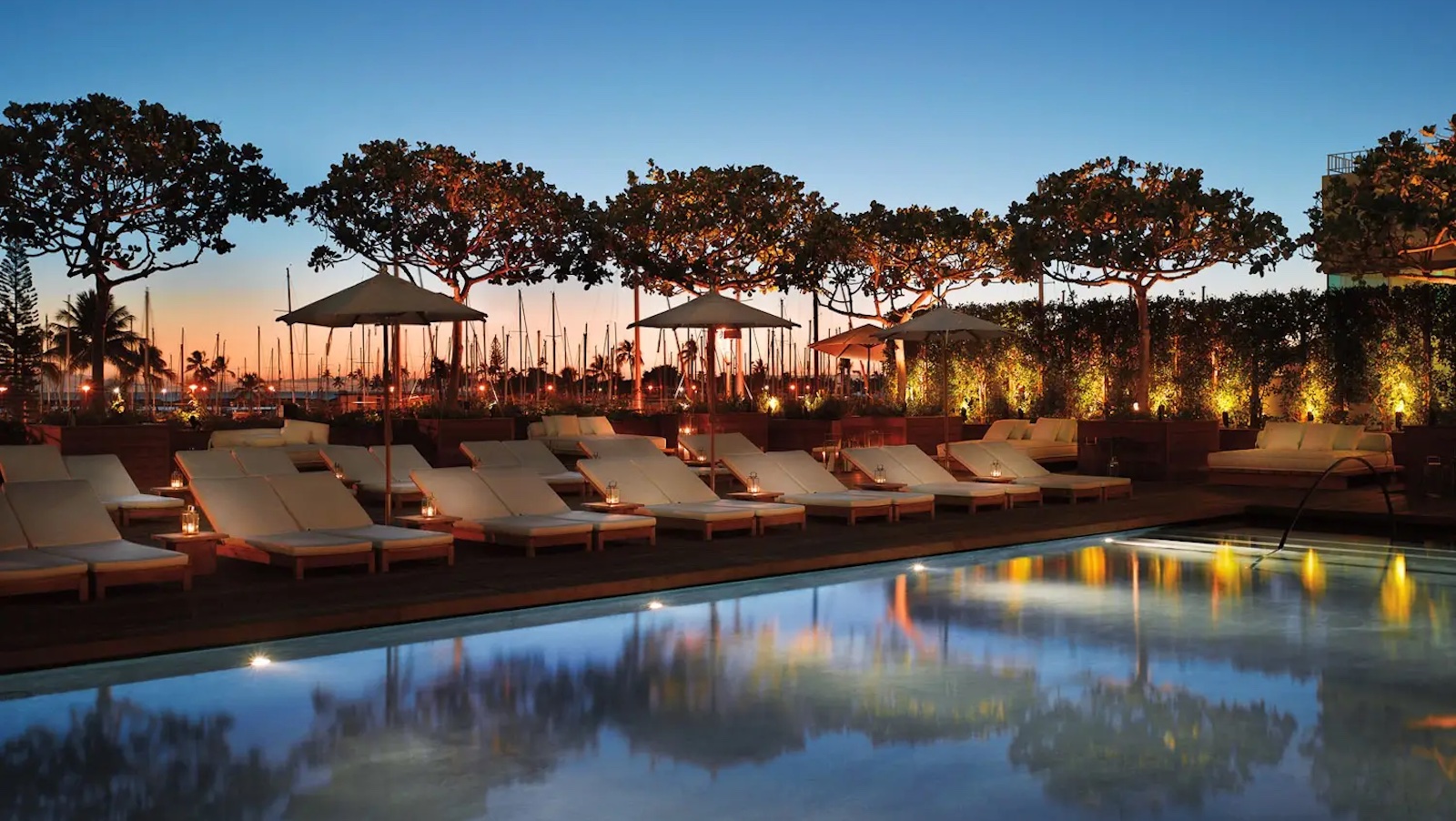 A serene poolside scene at sunset, featuring rows of white lounge chairs under umbrellas. The pool reflects the twilight sky, illuminated by warm lights lining the area. Tall trees and a marina with docked boats are visible in the background, perfect for a romantic getaway in Hawaii.