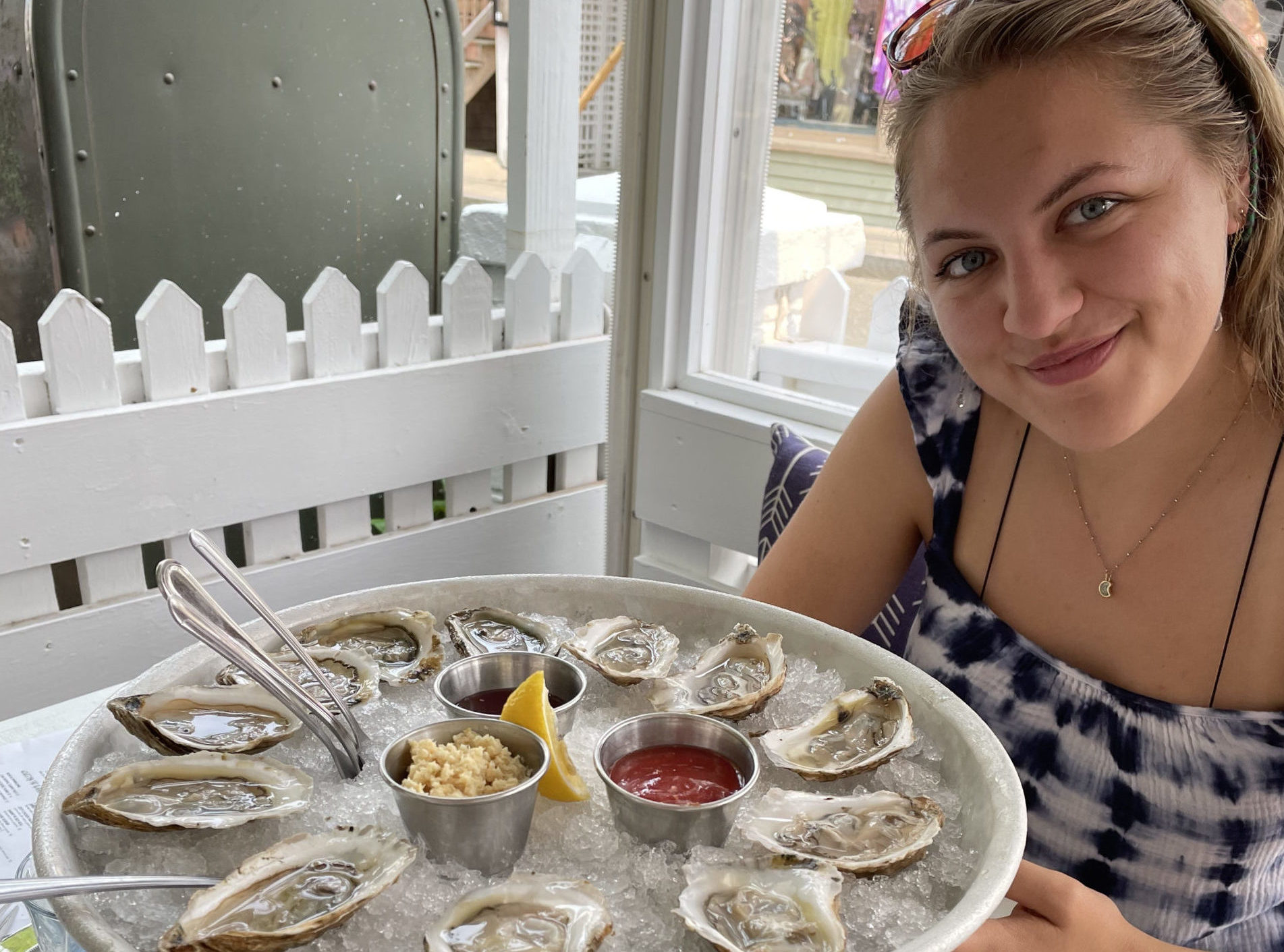 A smiling person is seated at a white table beside a white picket fence. In front of them, on the table, is a large platter filled with raw oysters on ice, lemon wedges, horseradish, and cocktail sauce. The setting appears to be a casual outdoor dining area.