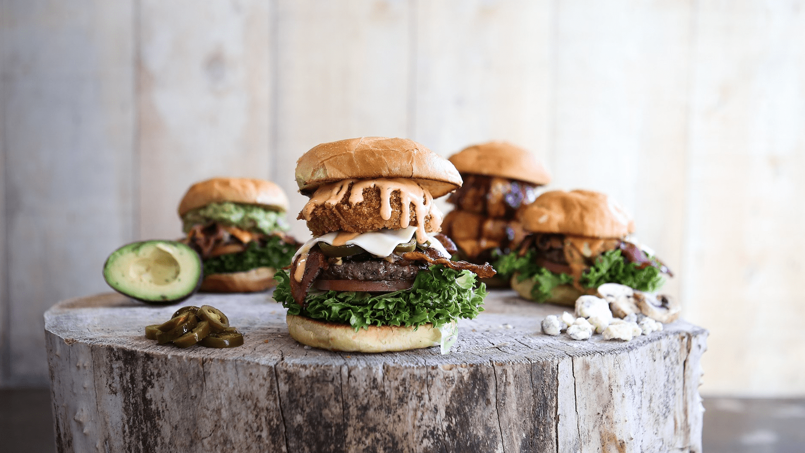 A rustic wooden table features a variety of gourmet burgers with different toppings, such as crispy onion rings, avocado, and jalapeños. Whole and sliced avocados and jalapeños are also on the table. The background is blurred, focusing attention on the burgers.