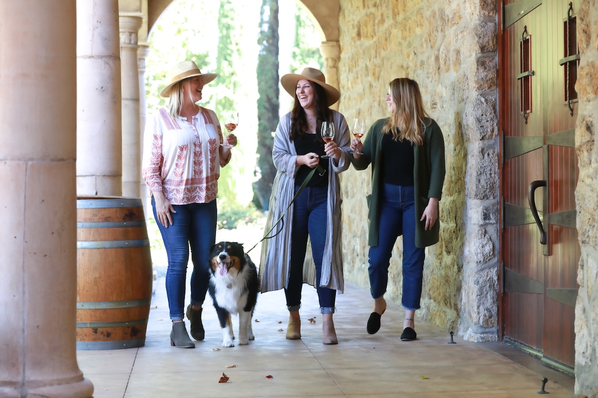 A group of three women walk under an awning on a walkway with a dog on a leash.