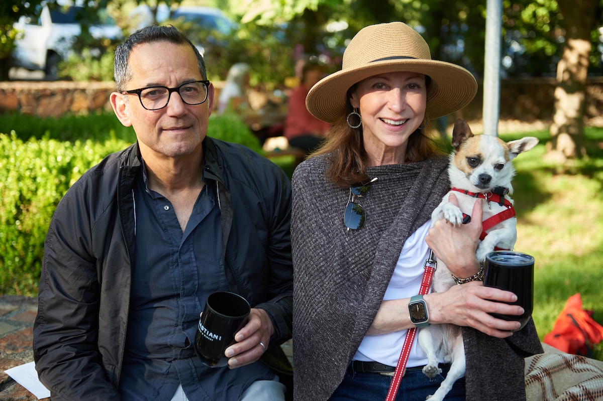 A woman wearing a sunhat and a man holding a cup sit with a small chihuahua.