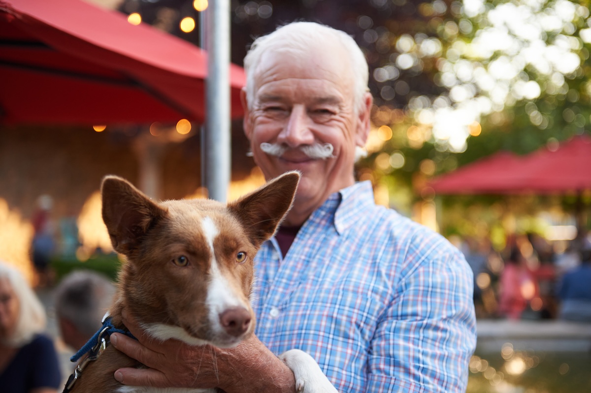 A man with a blue plaid shirt and a mustache holding his ginger-haired dog with white markings.