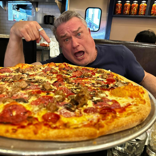 A man with a surprised expression points at a large pizza filled with various toppings, including pepperoni, sausage, and mushrooms. He is sitting in a restaurant booth, and jars of condiments are visible in the background on a shelf.