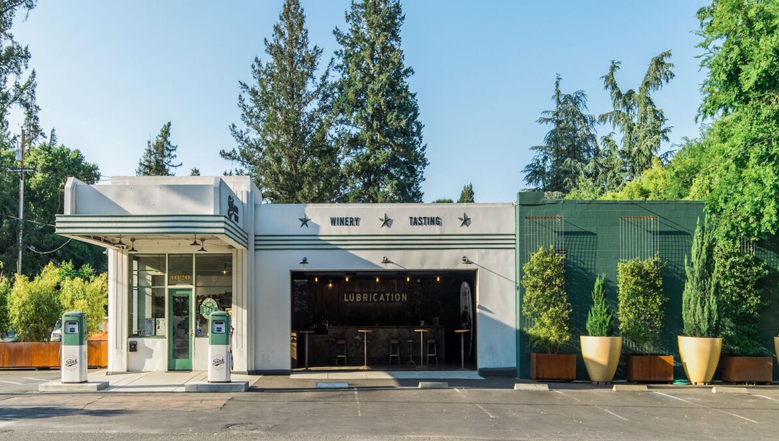 An exterior view of a building labeled "WINERY TASTING" with "LUBRICATION" on the door. It appears to be an old gas station repurposed into one of Napa Valley's charming tasting rooms. Two vintage gas pumps and lush green trees surround the dog-friendly area. The sky is clear.