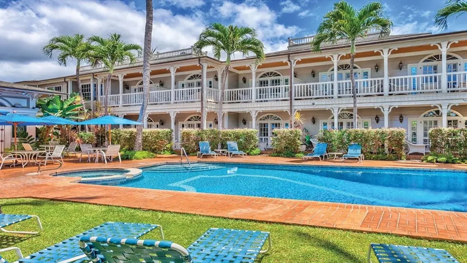 A romantic getaway awaits at this tropical Hawaiian resort, featuring a large blue swimming pool surrounded by brick paving and lounge chairs. The building behind has two stories with balconies, white railings, arch windows, and is decorated with palm trees and lush greenery. The sky is partly cloudy.