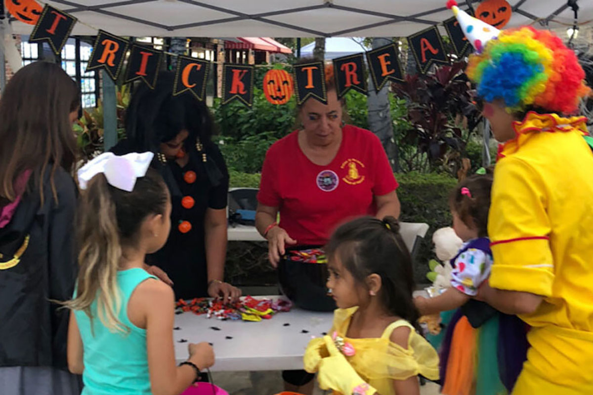 At Waikoloa Annual Events, a group of children in costumes gathers around a table under a canopy where adults are handing out candy. A clown and a woman in a red shirt distribute treats. Above the table, a "Trick or Treat" banner is displayed, decorated with pumpkins and bats.