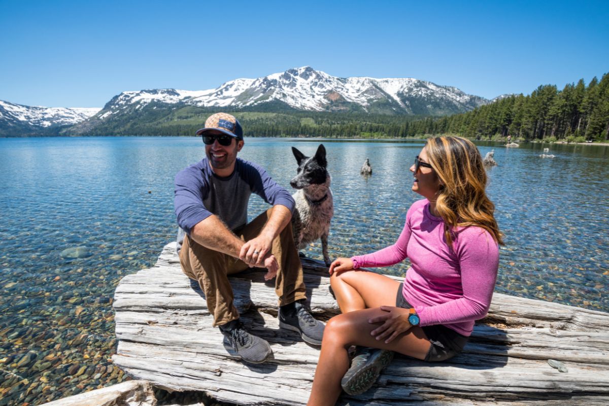 A man and a woman sit on a large log at the edge of pristine Lake Tahoe with a dog standing between them. Snow-capped mountains and pine trees grace the background under a clear, blue sky. The woman wears a pink top and shorts, while the man sports a blue top and shorts—perfect for their Dog Approved Hikes adventure.