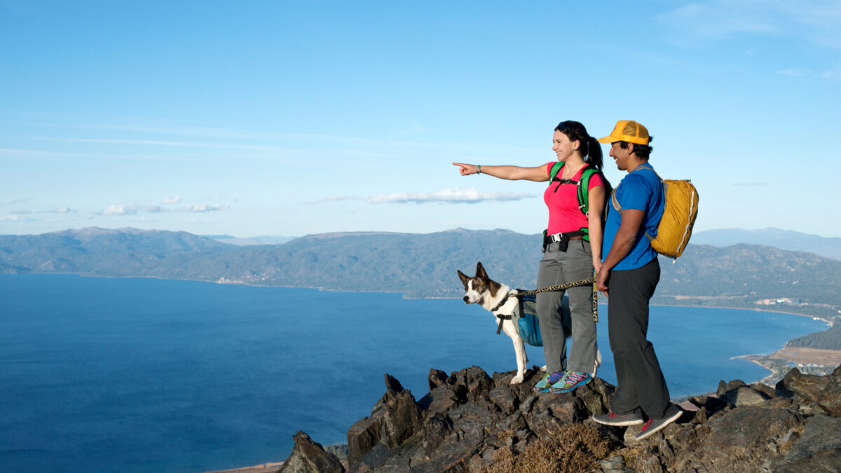 Two hikers and a dog stand on a rocky cliff overlooking Lake Tahoe and distant mountains. One hiker, wearing a pink shirt, points toward the horizon while the other in a yellow hat and blue shirt watches. The dog on a leash eagerly takes in the view, enjoying one of many Dog Approved Hikes.