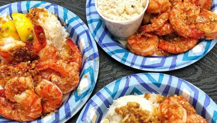 Three paper plates with a vibrant blue and white pattern, each holding garlic shrimp over white rice, are displayed on a dark wooden surface. A small cup filled with coleslaw is placed beside the topmost plate.