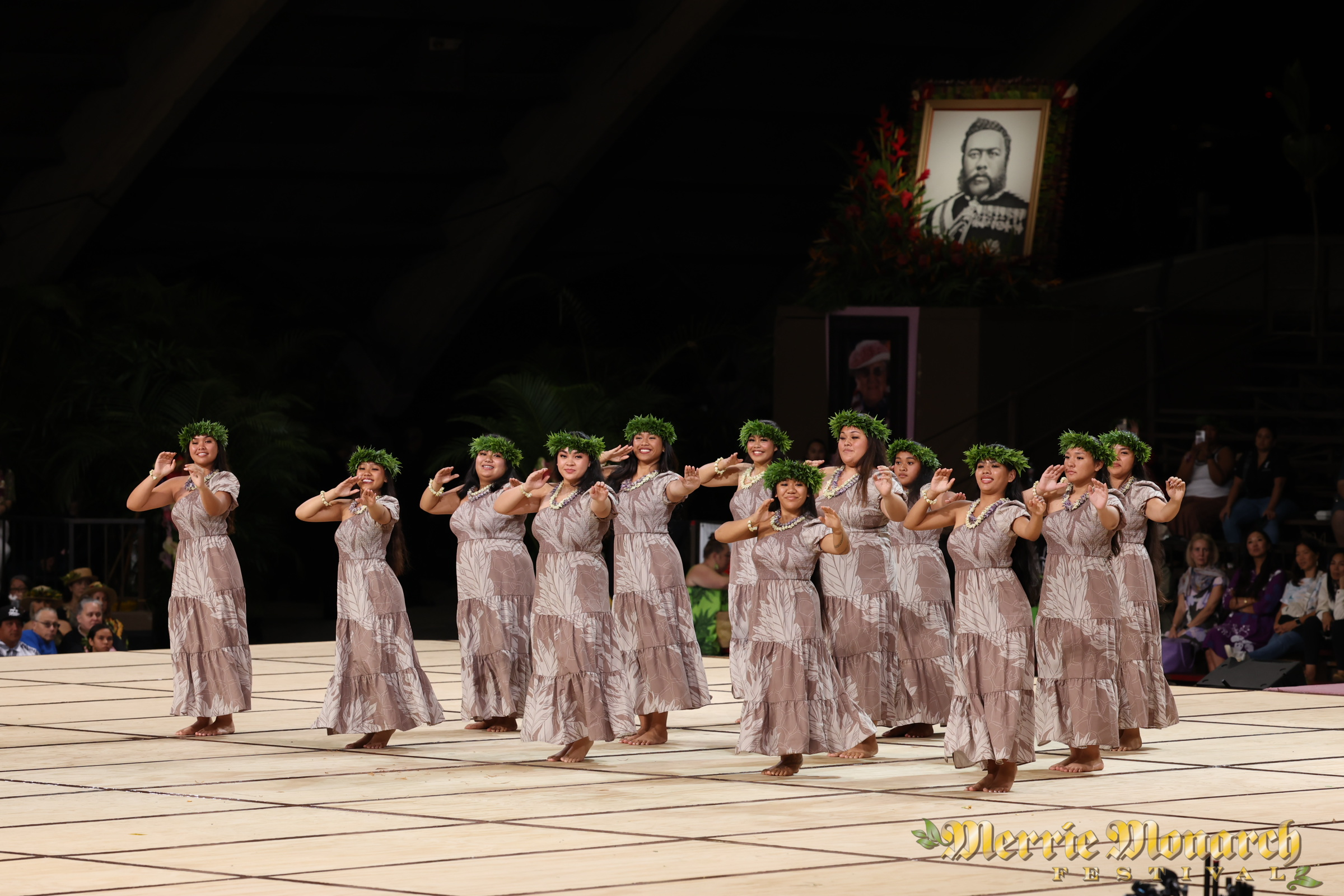 A group of hula dancers dressed in matching beige dresses and green leaf headpieces perform on stage at the Hilo Annual Events cultural celebration. A framed portrait of a historical figure is displayed in the background, with the stage captioned "Merrie Monarch" in the corner.