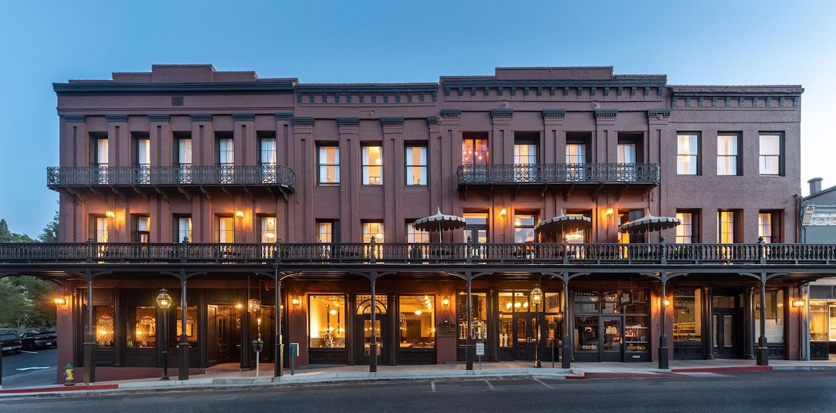 A large, historic, three-story brick building is set against a twilight sky. The facade of the National Exchange Hotel includes numerous windows and two levels of balconies with ornate railings. Warm lights glow from the interior, highlighting the building's vintage charm.