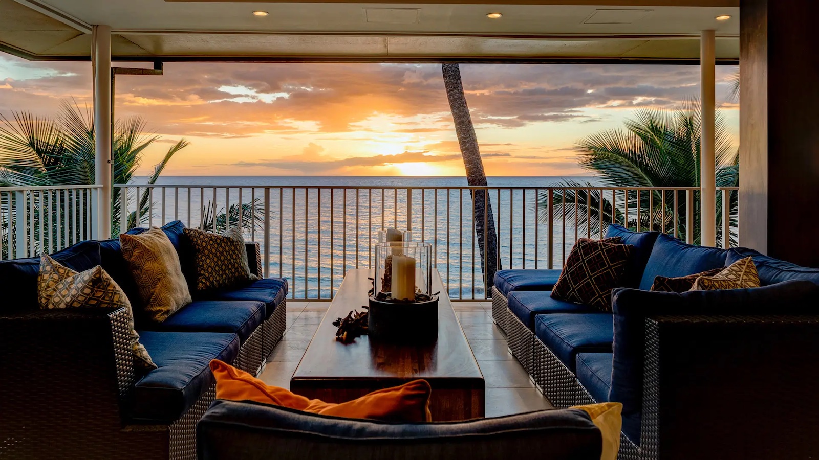 A cozy outdoor seating area with dark blue couches and a wooden coffee table overlooks a beautiful sunset over the ocean, creating the perfect luxury getaway in Hawaii. Palm trees frame the view, and warm light from the sunset creates a serene ambiance.