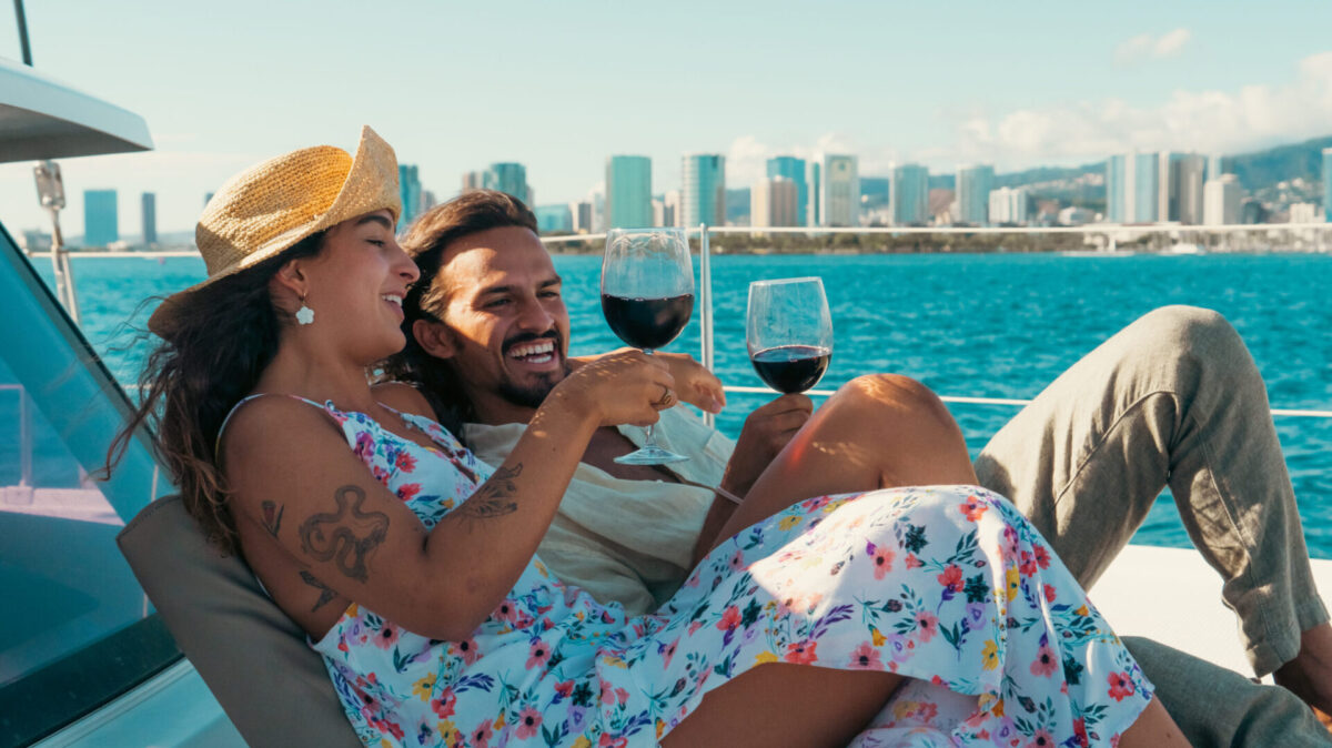 A couple lounges on a boat, laughing and enjoying glasses of red wine. The woman is wearing a floral dress and straw hat, while the man is casually dressed. The boat is on the water with a cityscape and mountains visible in the background under a clear sky.