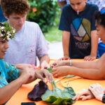 A group of people gathered around an orange table outdoors, engaged in a leaf weaving activity during the Waikoloa Annual Events. A woman wearing a floral crown is demonstrating the technique. Participants of various ages are attentively learning, surrounded by lush greenery.