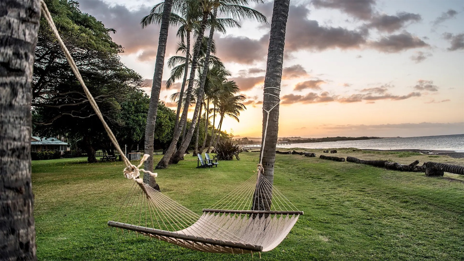 A tranquil beach scene at sunset, featuring a hammock tied between two palm trees on grassy terrain. In the distance, other palm trees line the beach with a few chairs set up near the shore. The sky is painted with light clouds and warm hues, reminiscent of value hotels in the Hawaiian Islands.