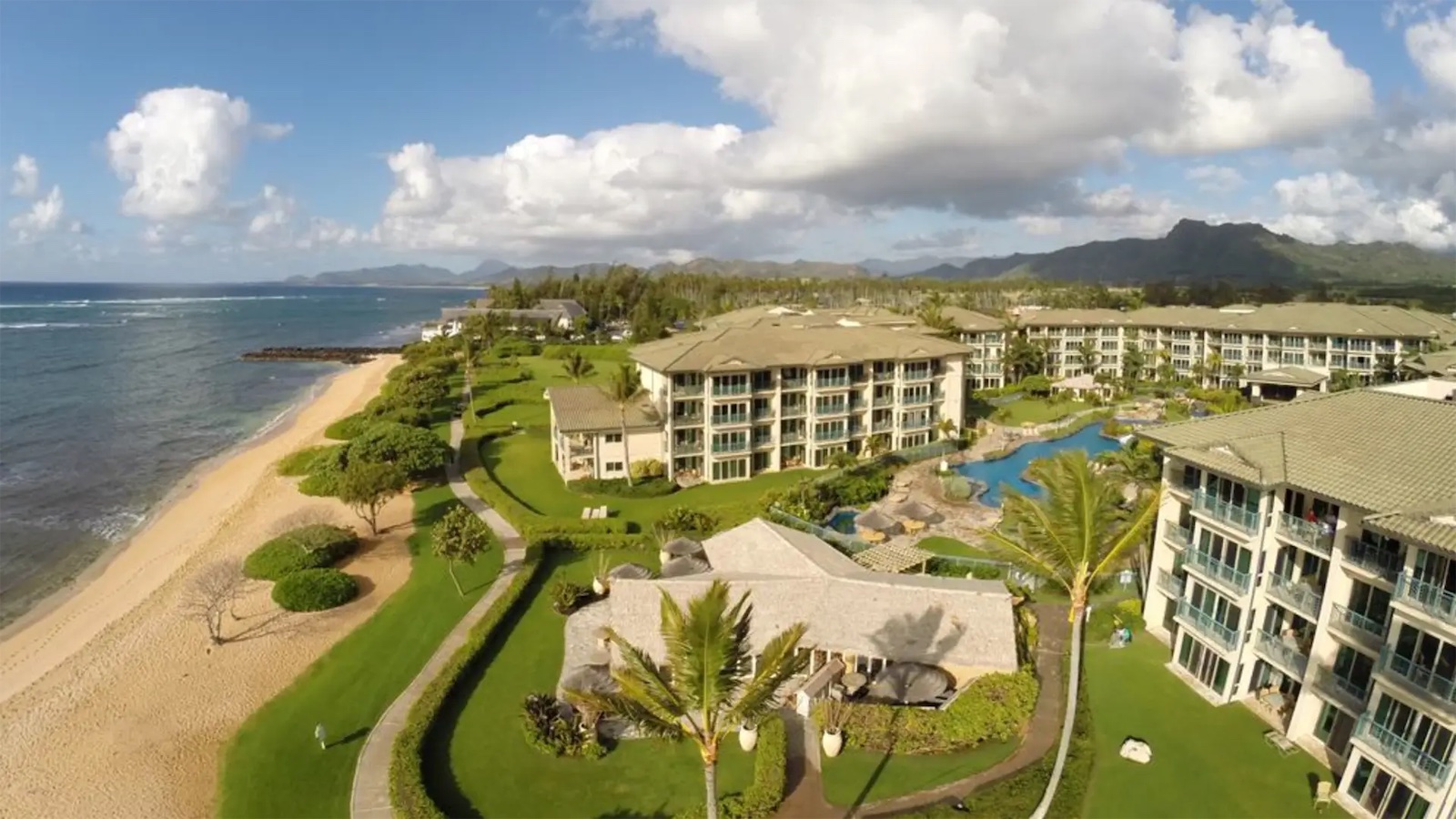 Aerial view of a beachfront resort featuring multiple multi-story buildings with balconies, a large pool, and lush green landscaping. The resort is adjacent to a sandy beach with gentle waves, set against a backdrop of mountains and partly cloudy skies.