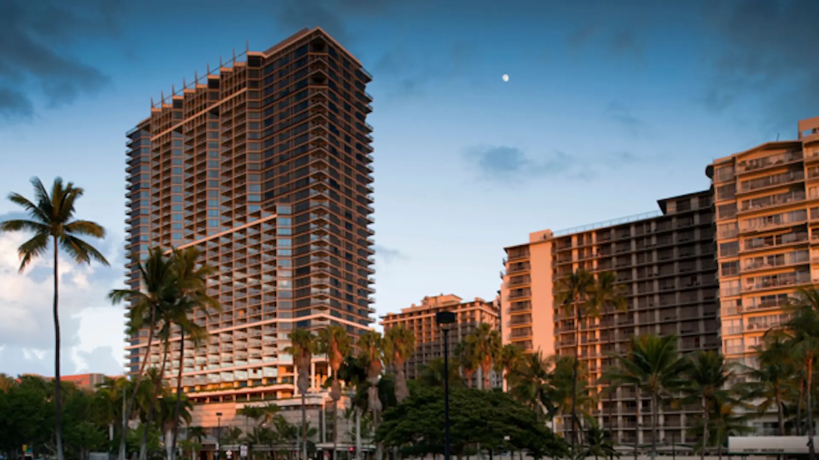 A group of modern high-rise buildings stand tall, illuminated by the warm light of the setting sun. Palm trees line the foreground, contrasting with the urban architecture. The sky is clear with a few clouds and a small moon is visible—a perfect luxury getaway reminiscent of Hawaii.