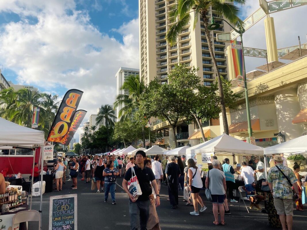 People walk around in the streets for the Kalakaua Festival visiting booths, white a view of the sky and city is in the background.