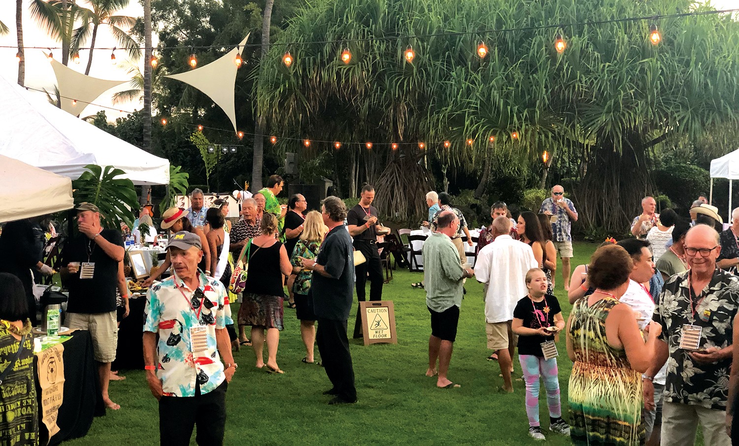 Guests mill around outside on a grassy area with lights and booths set up at the Kona Cultural Coffee Festival.