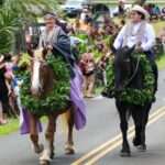 There are two people on horseback with leis riding through the streets of South Maui for the Festivals of Aloha, with a crowd watching.