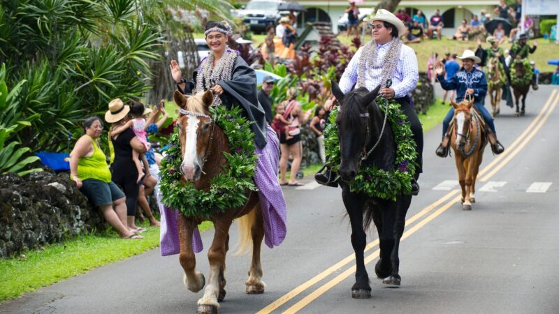 There are two people on horseback with leis riding through the streets of South Maui for the Festivals of Aloha, with a crowd watching.