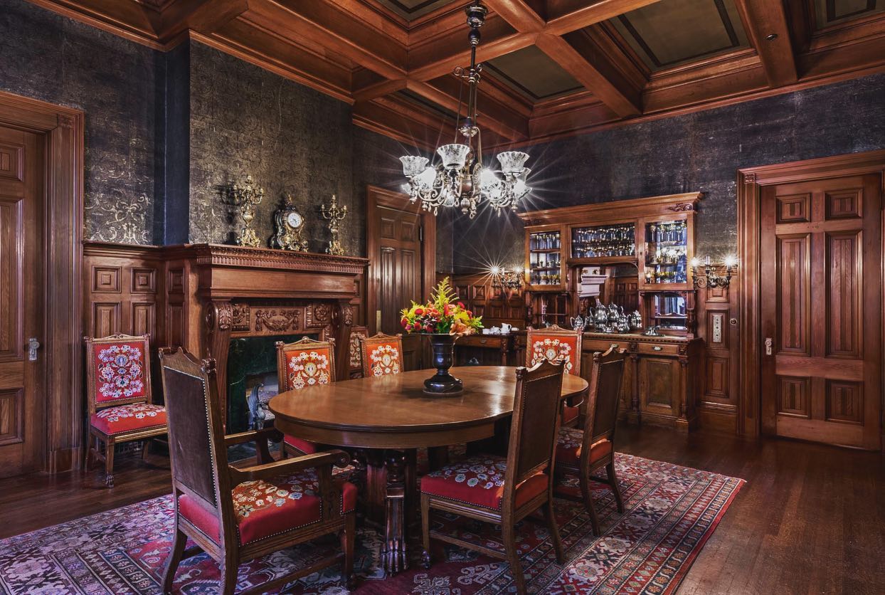 Photo of a dining room with lots of dark wood on the walls, ceilings, floors and furniture. There is a dining table and chairs with a chandelier above, fireplace and cabinetry.