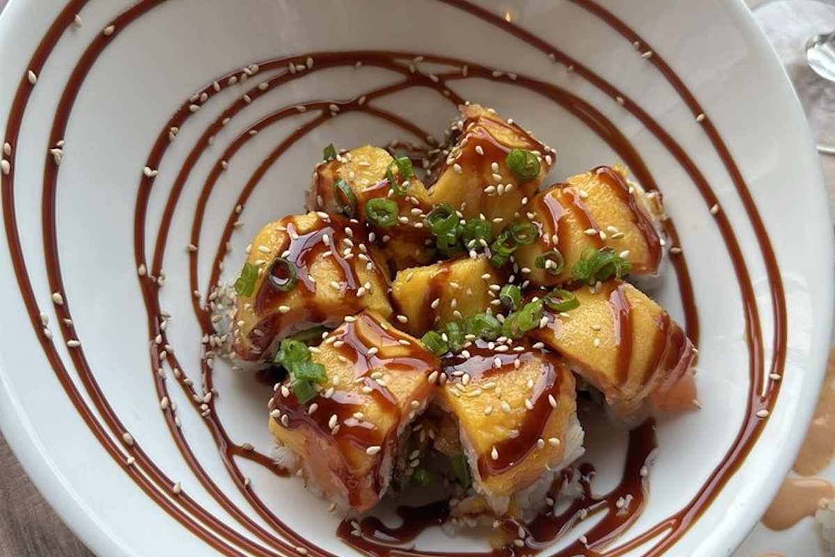 A bowl of sushi artfully arranged with sesame seeds, green onions and a swirled brown sauce on top.