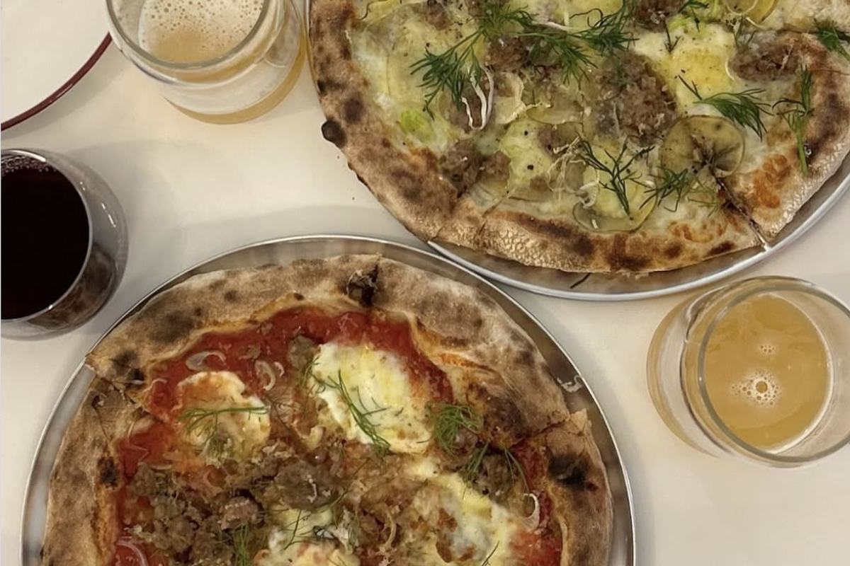 Two pizzas with lots of toppings and herbs and beverage glasses on the side.