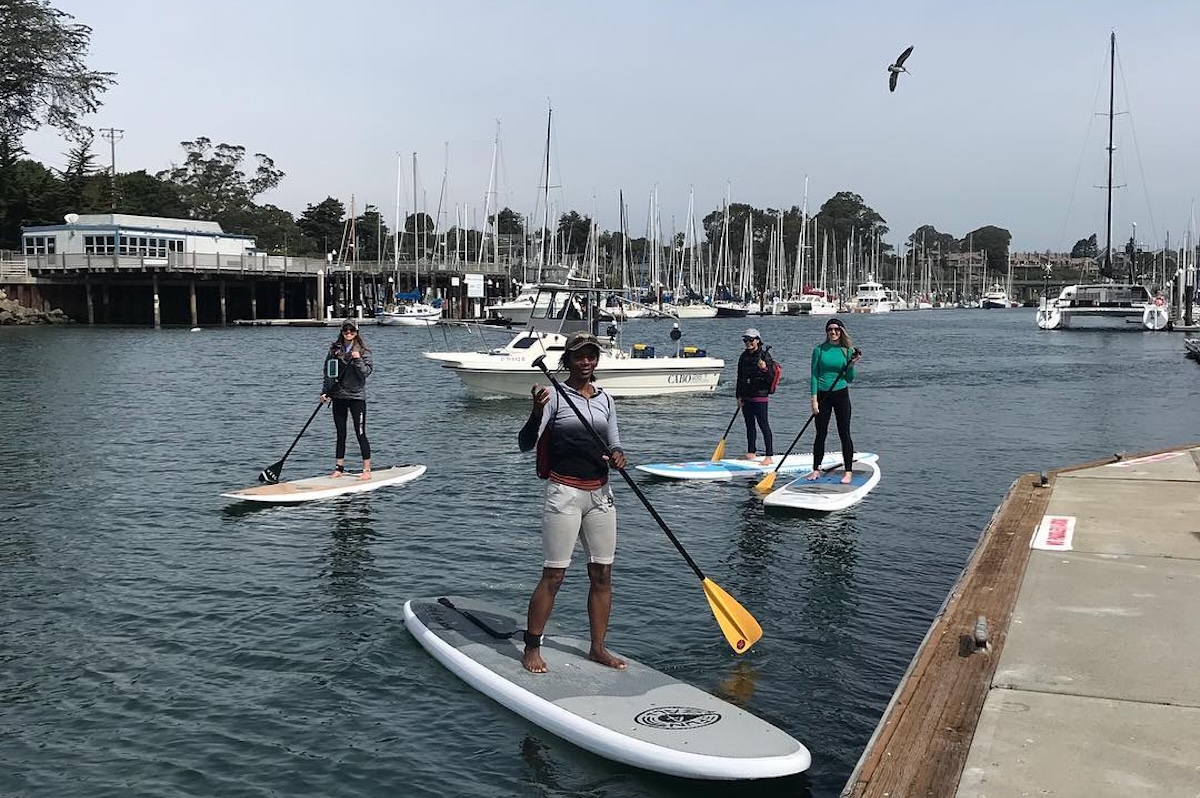 Four people stand on standup paddleboards holding oars near a dock and with boats in the background.