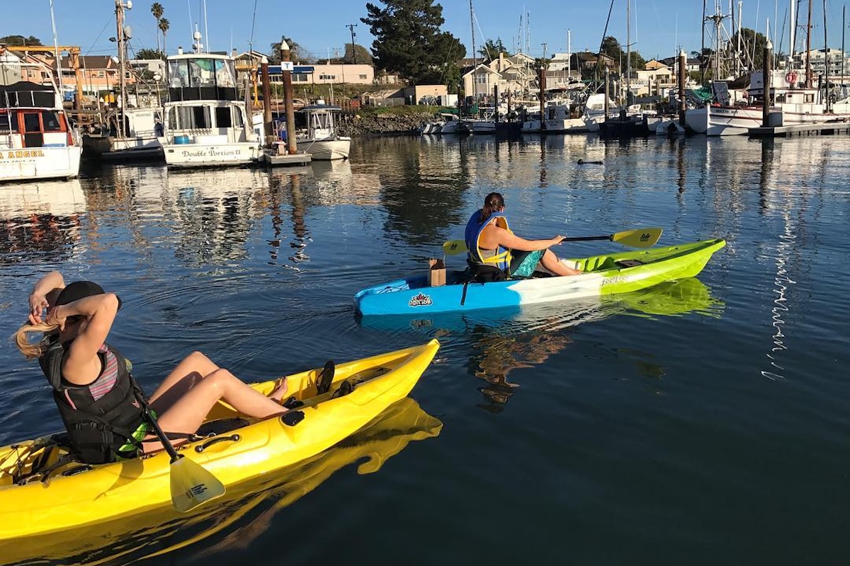 Two people sit in kayaks in the bay with boats in the background.