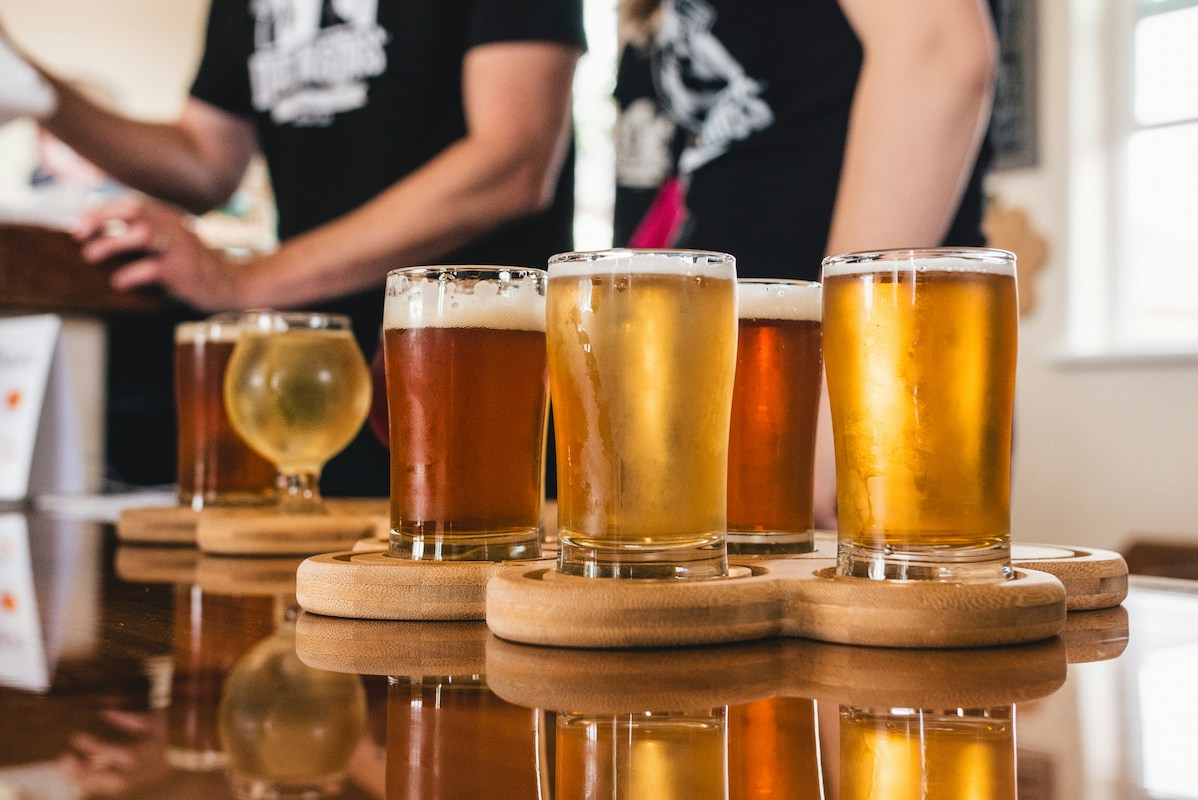 Cups filled with beer sit on coasters on a counter with two people standing in the background.