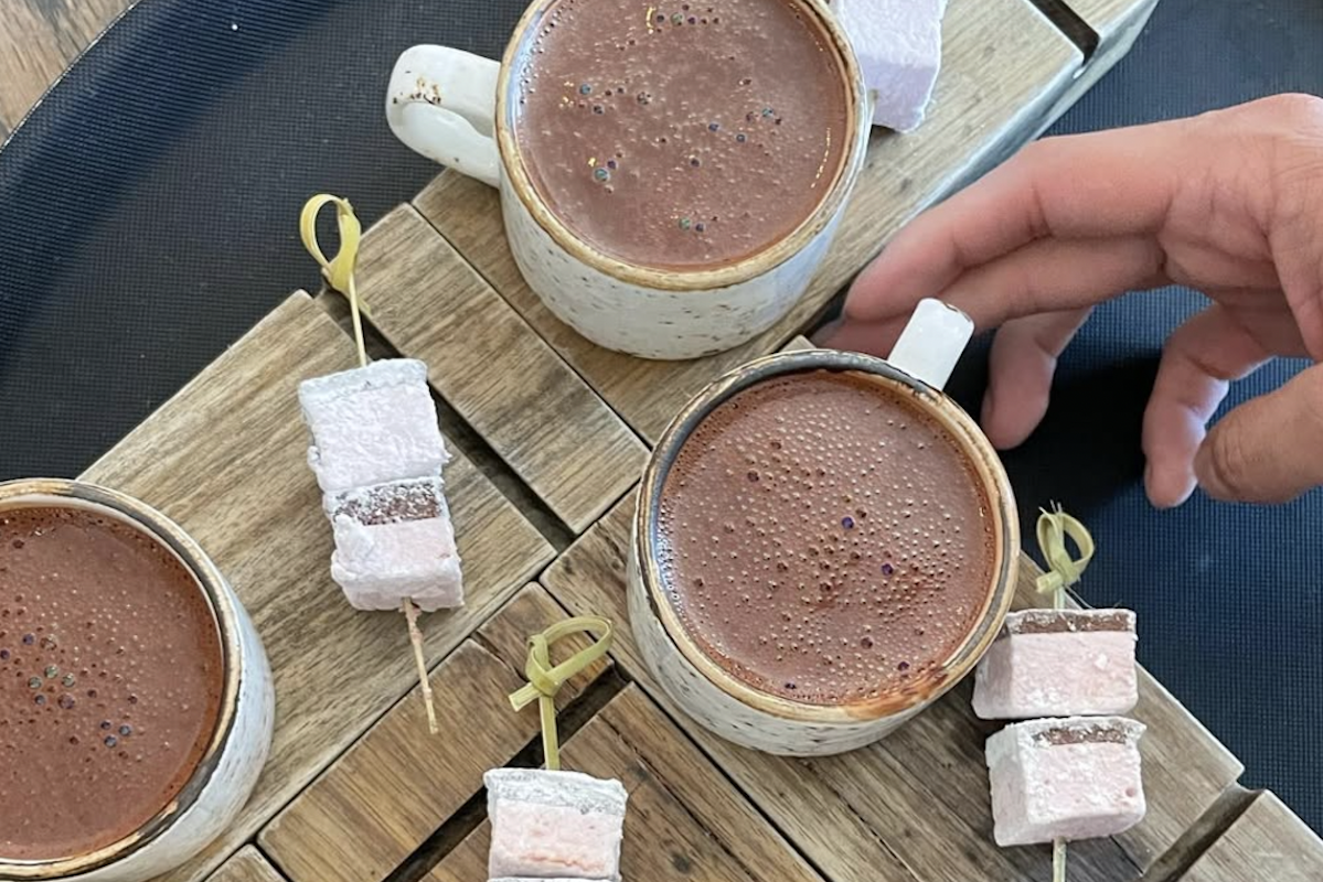 Cups of a dark brown liquid with marshmallows on skewers sit on a wooden platform as someone reaches for one of the mugs.