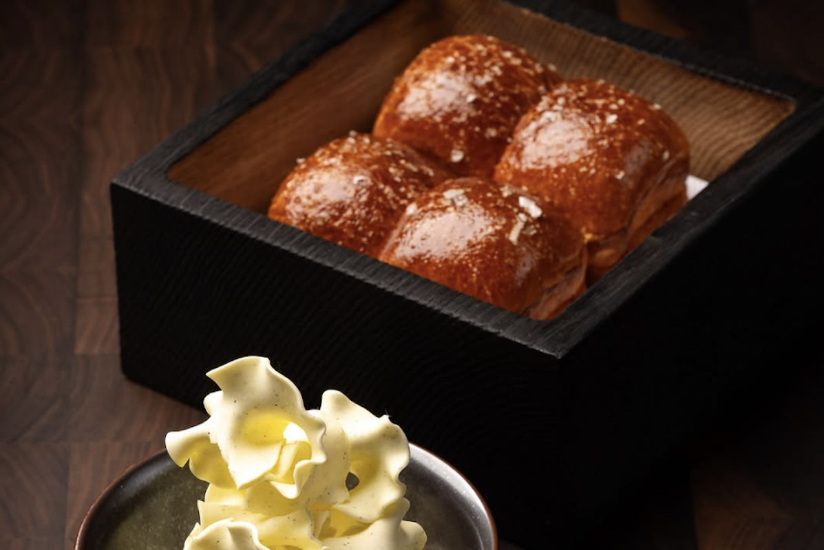 Four glossy bread rolls sit in a black box container with butter sculpted to look like seaweed.