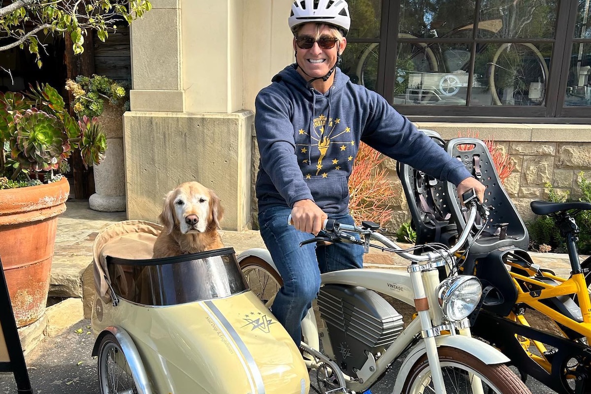 A man on an electric bike with a dog in a side car poses in front of a building.