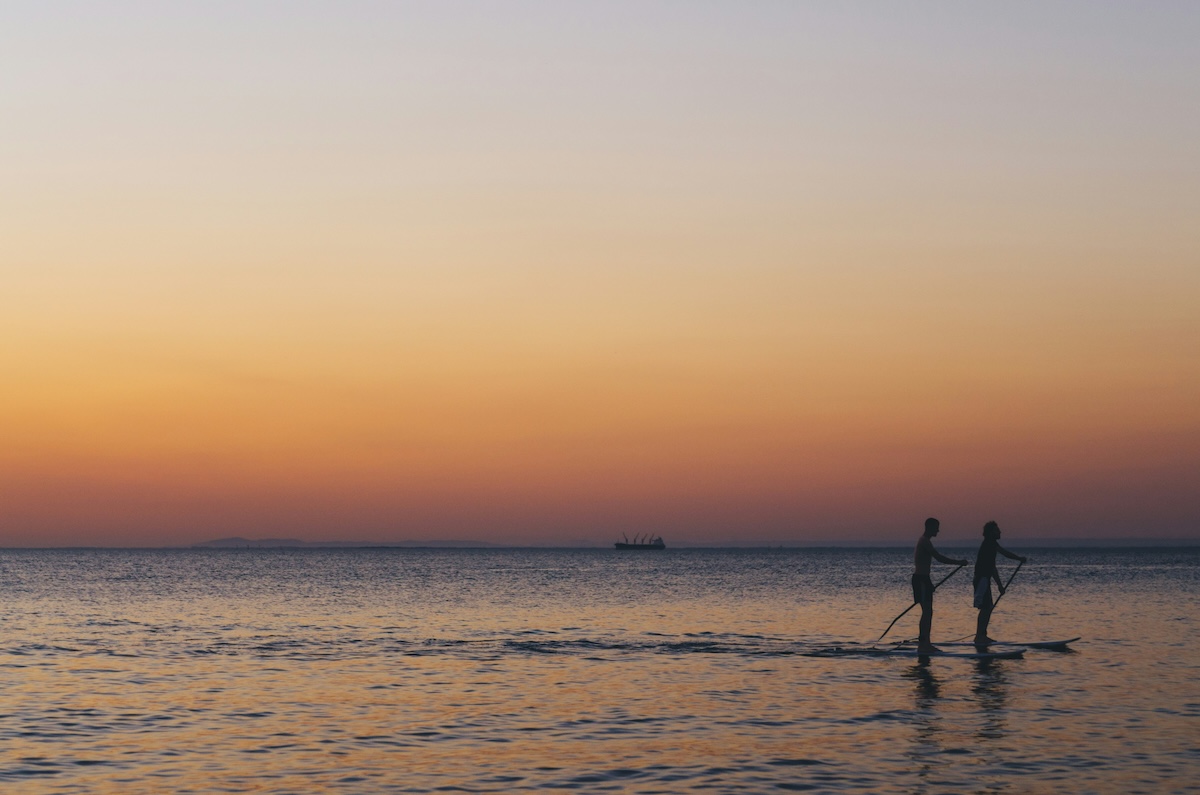 Two silhouettes paddleboard on the ocean while the sun sets in the background.