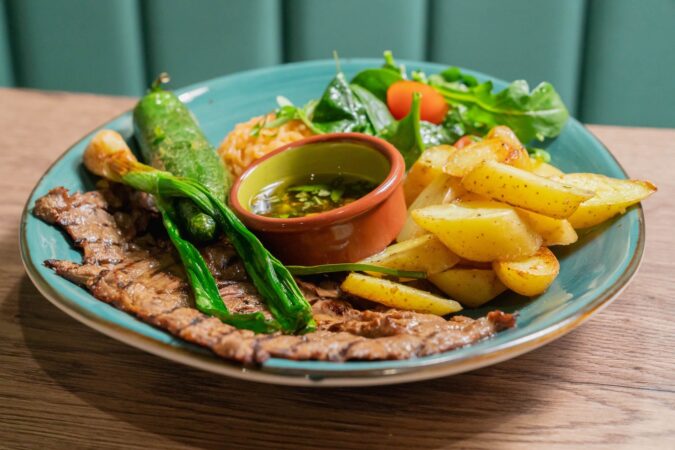 A greenish blue plate with some sort of flattened grilled meat, potatoes, salad, green veggie and a tiny bowl of sauce.