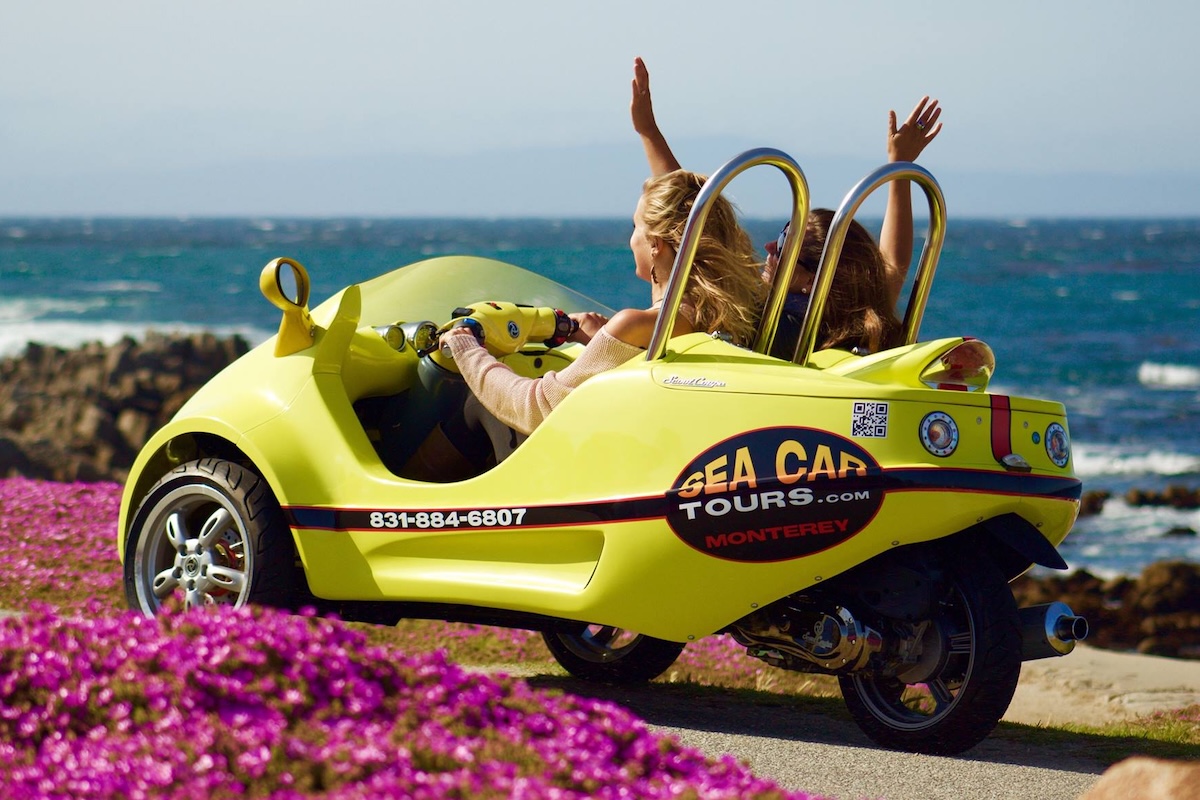 Two girls drive in a yellow GoCar surrounded by purple flowers and the ocean view.