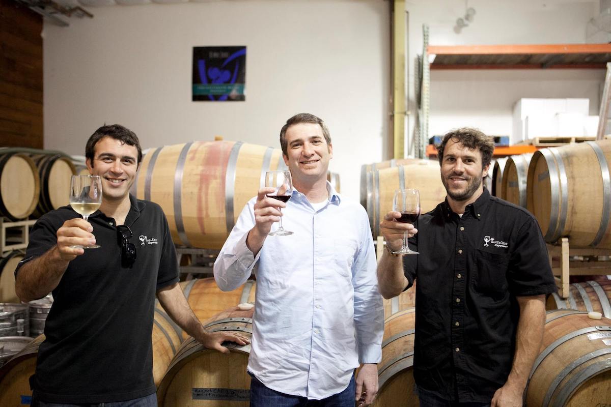 Three men hold glasses of wine standing in from of wine barrels.