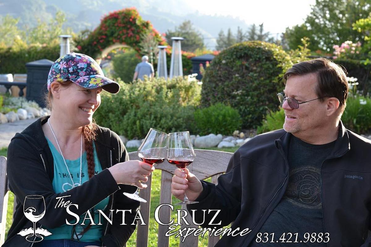 Two people cheers wine glasses on a bench on a grassy area.