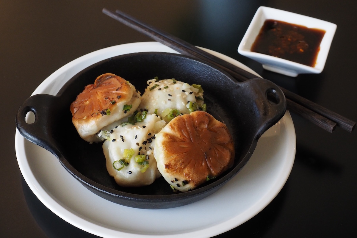 A small cast iron pan holds four sheng jian bao, a type of dim sum, with green onions and black sesame seeds on top and chopsticks and dipping sauce on the side.