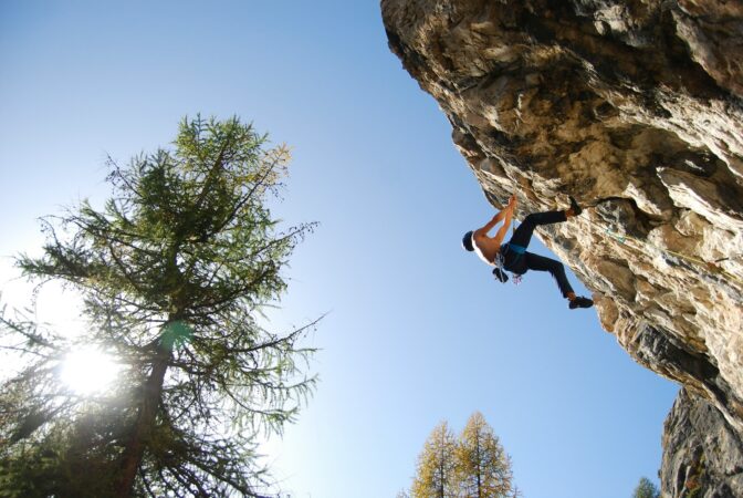 A man dangles high up on the side of a rocky edge, as he rock climbs against a clear blue sky.