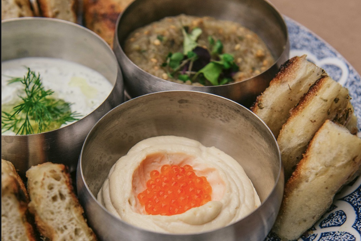 A bowl of hummus with roe and two other dipping sauces garnished with herbs and breads on the side.