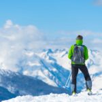 A man wearing a lime green jacket and a backpack holds hiking poles on top of a snowy mountain overlooking other peaks.